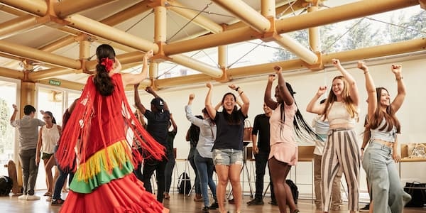 A diverse group of people are joyfully participating in a dance class led by a woman in a colorful traditional dress.