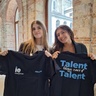 Two women smiling and holding up black T-shirts with 'Talent taking care of Talent' text in a historical building.