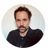 A man with a slight beard and tousled hair looks directly at the camera against a plain background.