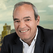 A portrait of a smiling middle-aged man wearing a black suit and white shirt, with a cityscape in the background.