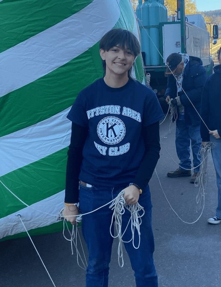 A young person holding ropes in front of a large green and white striped tent, wearing a black t-shirt with 'Preston Area Kiwanis Club' logo.