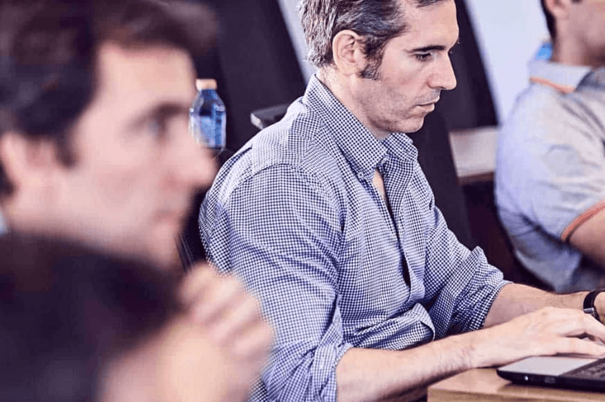 A professional man working on a laptop in a conference room setting with blurred colleagues in the background.