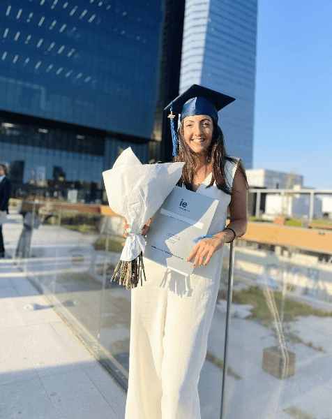A female graduate holding a bouquet and her diploma in front of a modern building.