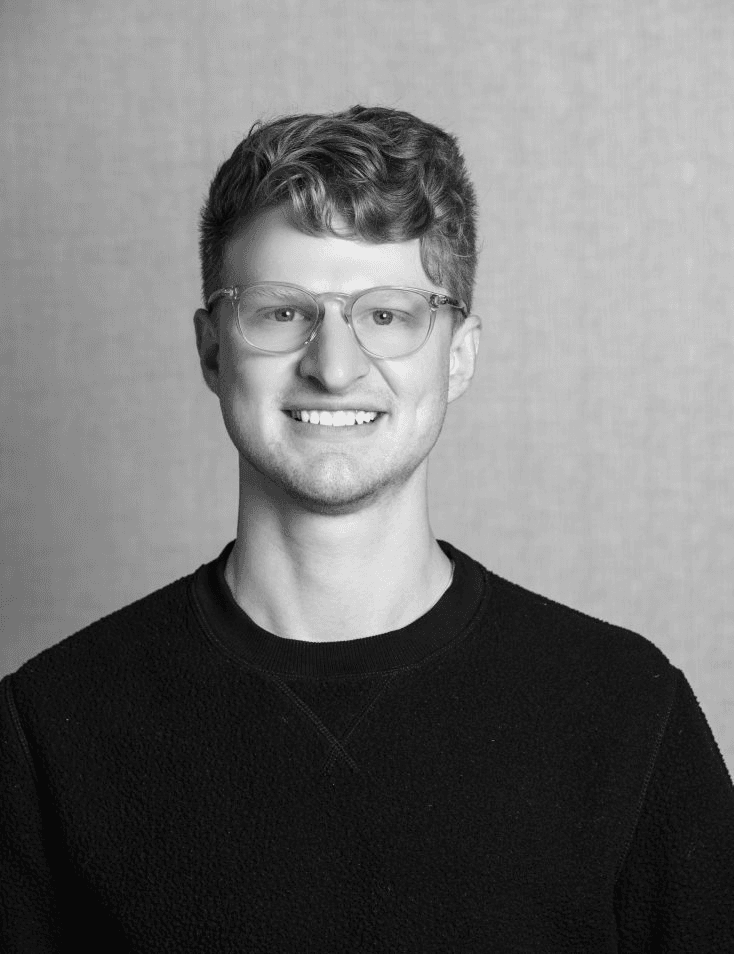 Black and white portrait of a smiling young man with glasses and curly hair wearing a black sweater.