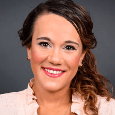 A portrait of a smiling woman with curly hair, wearing a light pink blouse and subtle makeup.