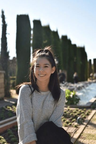 A young woman smiling in a sunny park with trees and a fountain in the background