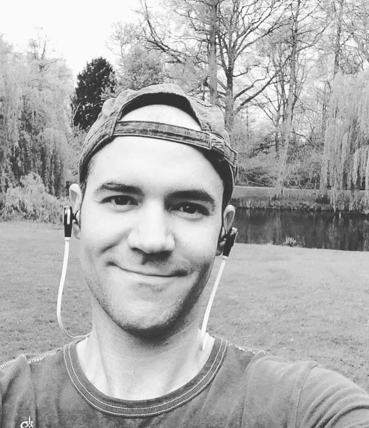 A smiling man wearing a baseball cap and earphones in a park setting with trees and a pond in the background.