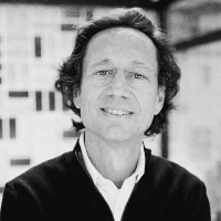 Black and white photo of a smiling middle-aged man with long hair, standing indoors with blurred buildings in the background.