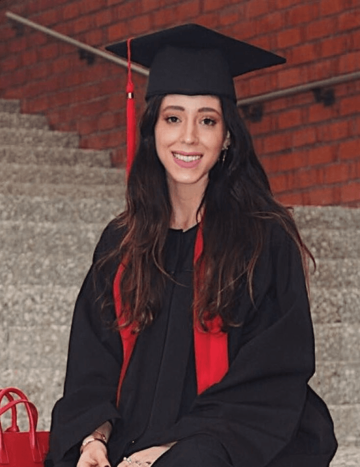 A young woman in graduation attire sitting on steps and smiling at the camera.