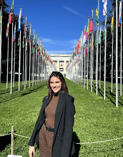 A woman smiling in front of a row of international flags outside a building.