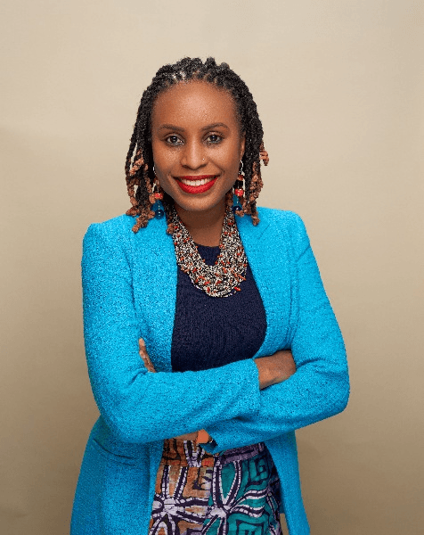 A confident woman in a blue blazer and colorful necklace standing with crossed arms.