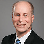 A professional headshot of a smiling bald man wearing a black suit and a blue striped tie.