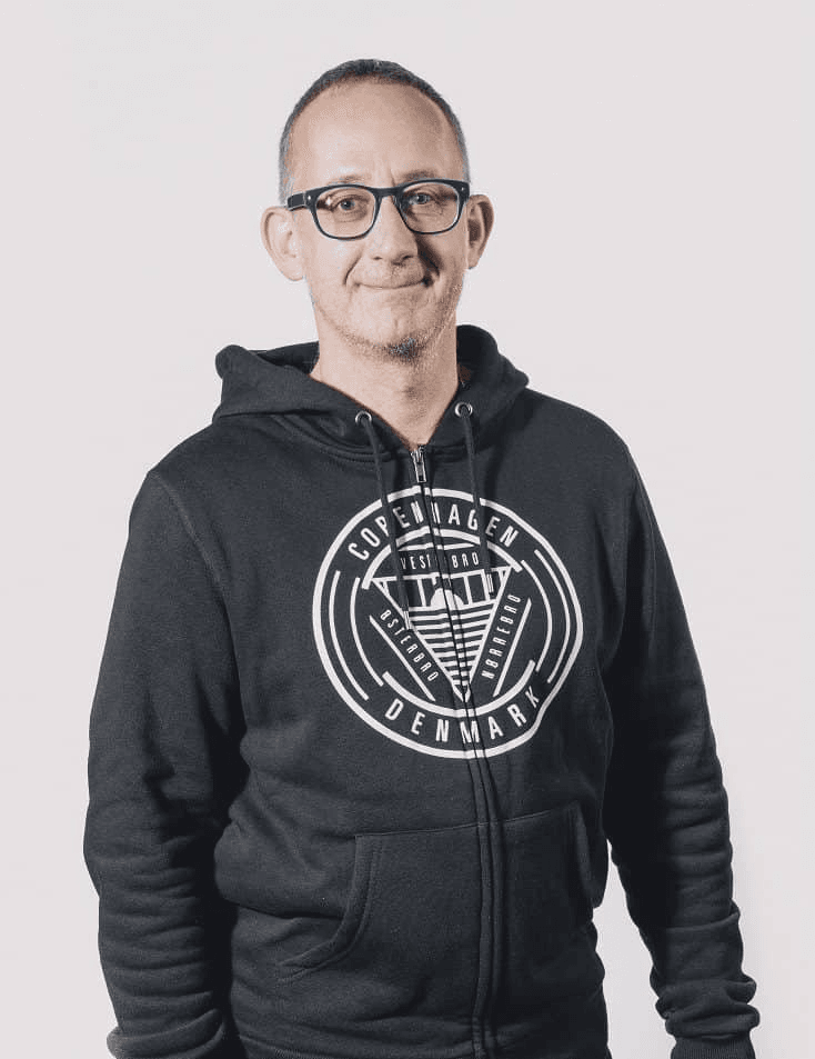 A middle-aged man wearing glasses and a black hoodie with 'Copenhagen, Denmark' printed on it, stands against a light grey background.
