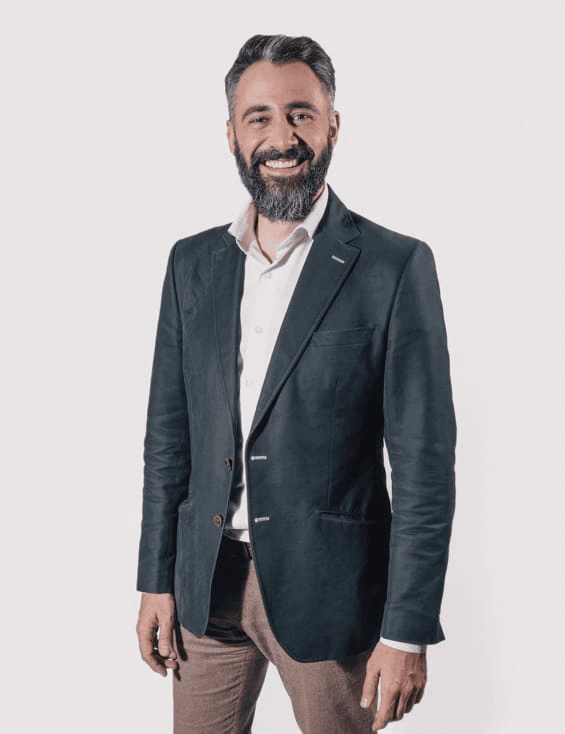A smiling man with a beard, wearing a black blazer and brown trousers, standing against a white background.