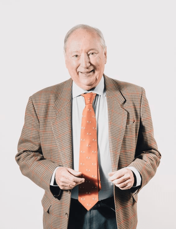 An elderly man in a patterned blazer and orange tie smiling at the camera.