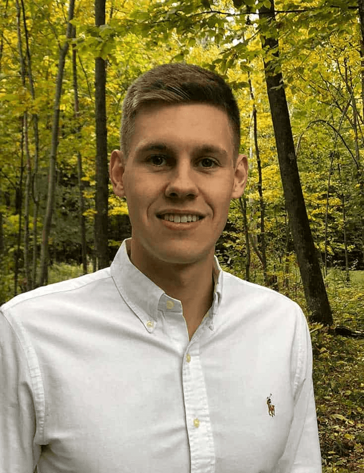 A young man in a white shirt smiling in a lush forest during autumn.