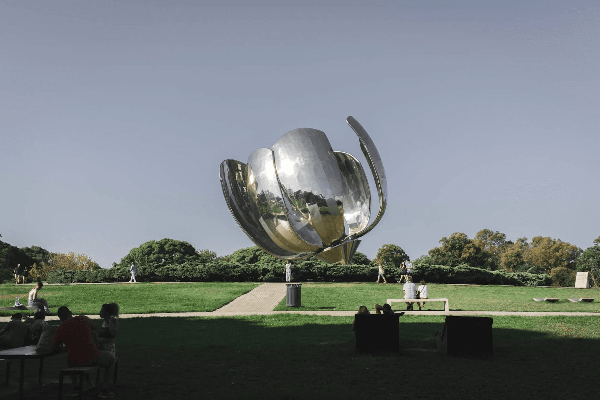 A large metallic sculpture resembling a flower stands at the center of a park, surrounded by people relaxing and enjoying the scenery.