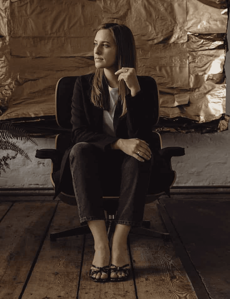 A woman in a thoughtful pose sits on a chair, dressed in a dark blazer and jeans, against a rustic backdrop.