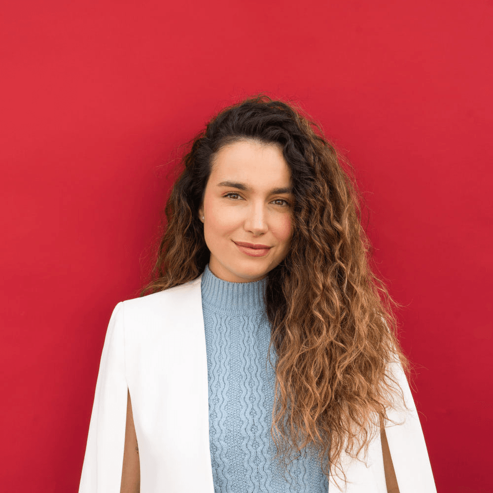 A young woman with curly hair, wearing a white jacket and blue turtleneck, stands in front of a red background.