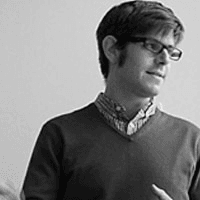 A black and white photo of a man wearing glasses, a sweater over a shirt, and looking to his left with a slight smile.