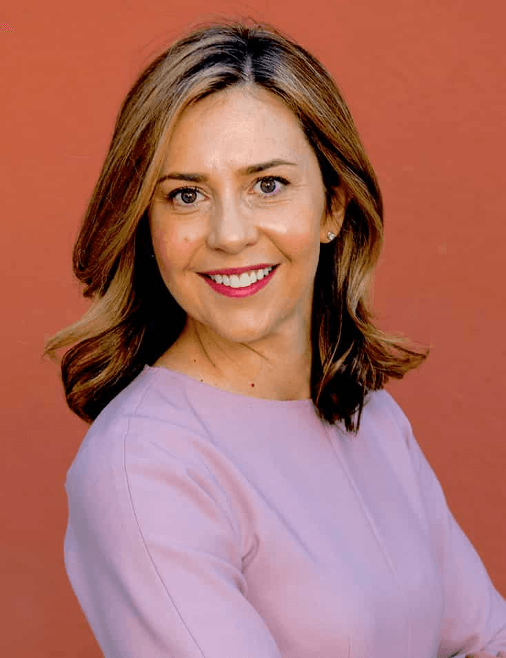 A smiling woman with medium-length hair, wearing a pink blouse, standing against a red background.