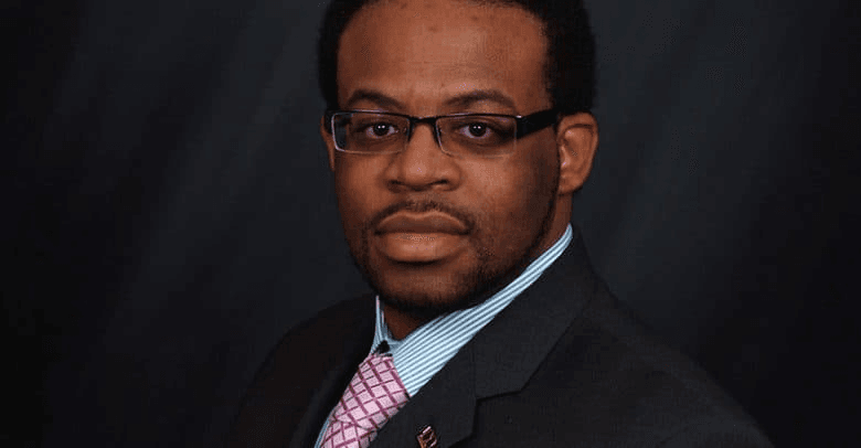 Portrait of a man wearing glasses, a blue blazer, and a pink tie.