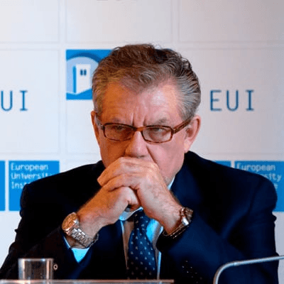 An elderly man with glasses is sitting at a desk with folded hands, looking thoughtful in front of a microphone and a backdrop that reads EUI.