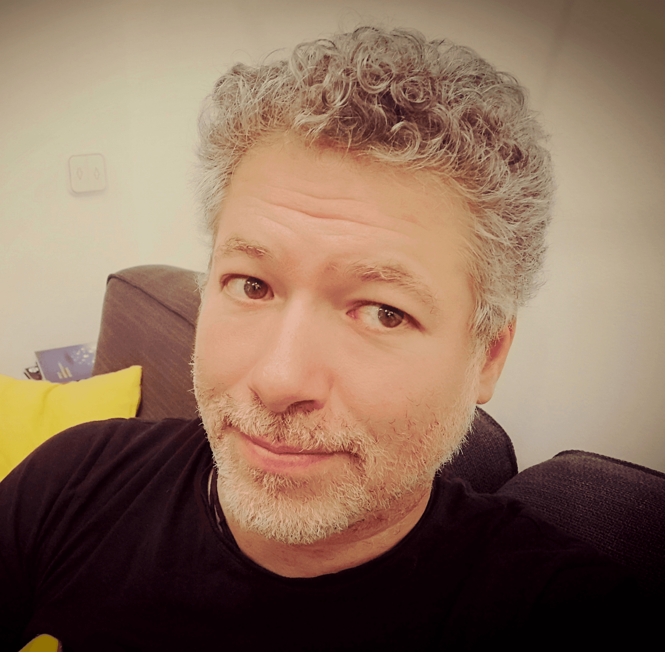 A close-up portrait of a middle-aged man with curly gray hair and a slight smile, sitting in a room with a yellow pillow and blue sofa in the background.