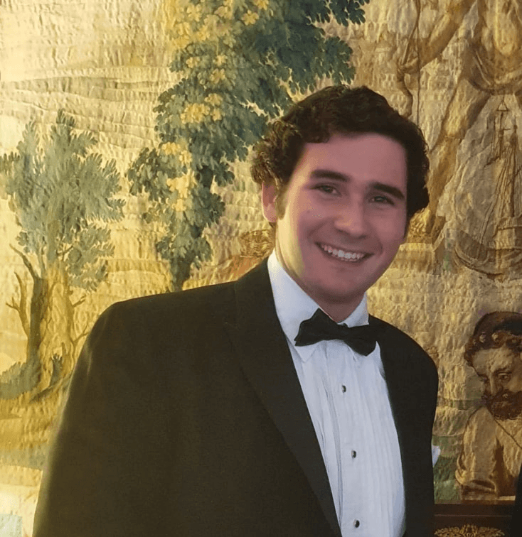 A young man in a tuxedo smiling in front of a decorative tapestry.