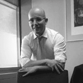 A black and white photo of a smiling bald man in formal attire leaning on a chair.