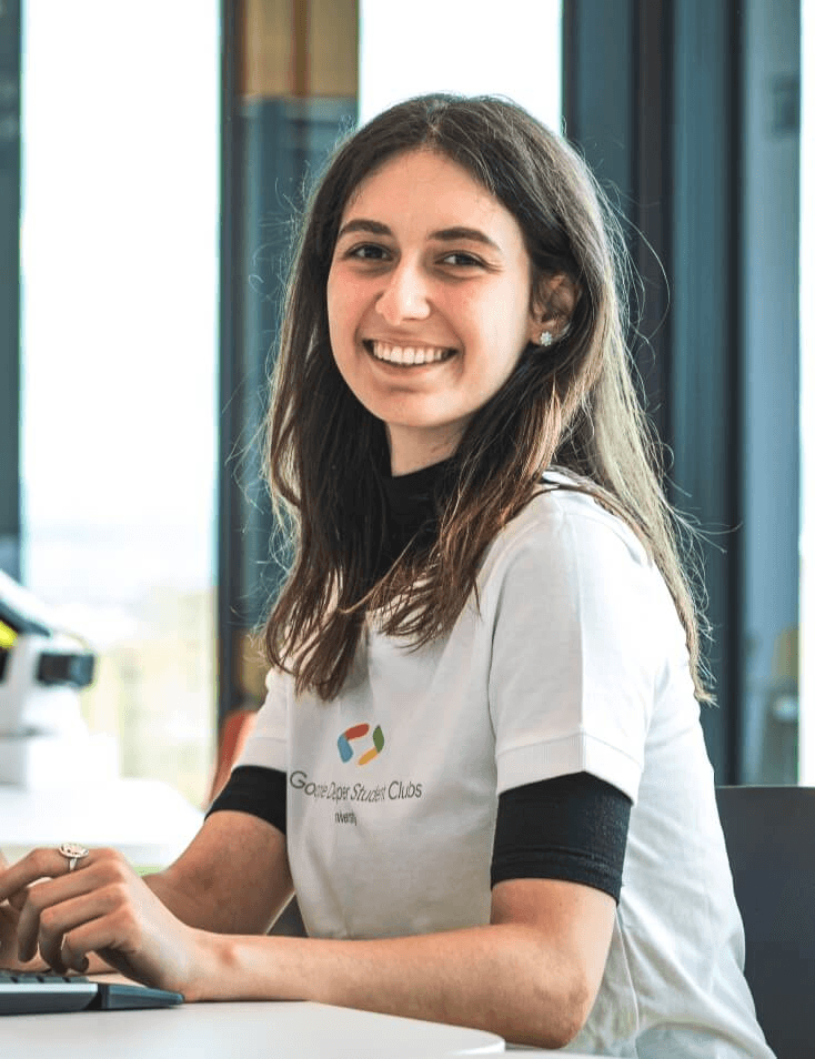 A young woman smiling at the camera while sitting at a desk with a laptop, wearing a white t-shirt with a 'Google Developer Clubs' logo.