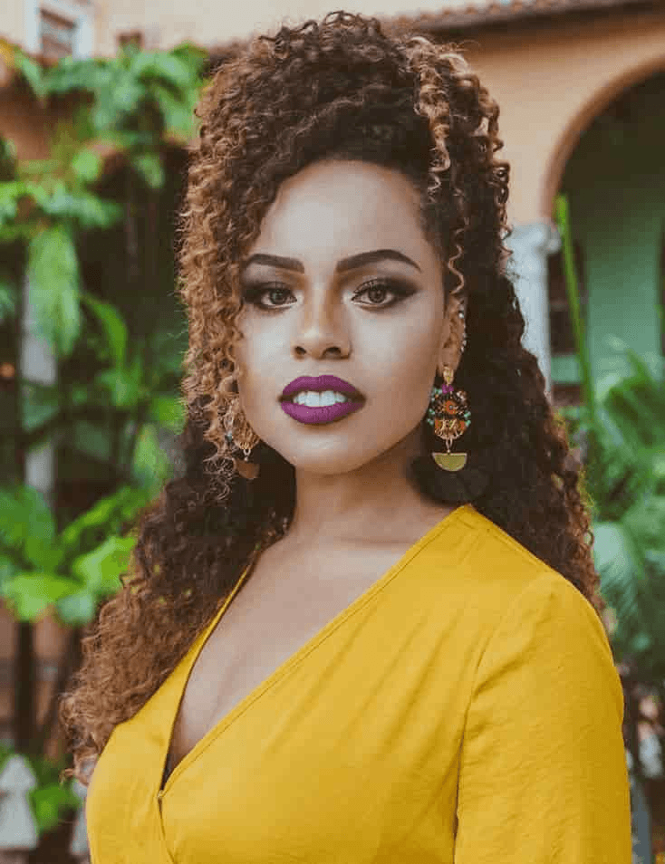 A portrait of a woman with curly hair wearing a yellow dress and bold makeup.