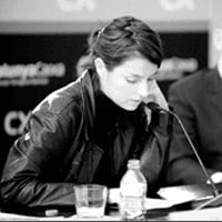 A woman in a black jacket is speaking into a microphone at a table with bottled water visible.