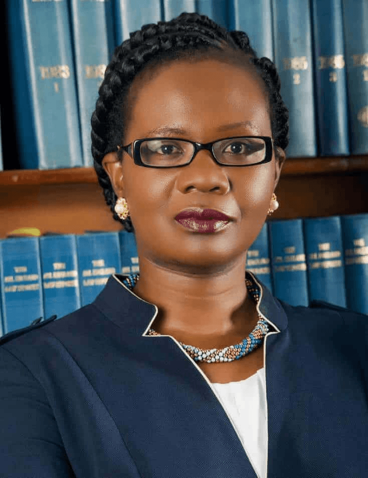 A professional portrait of a woman with braided hair, wearing glasses and a dark blue blazer, standing in front of a bookshelf filled with blue books.