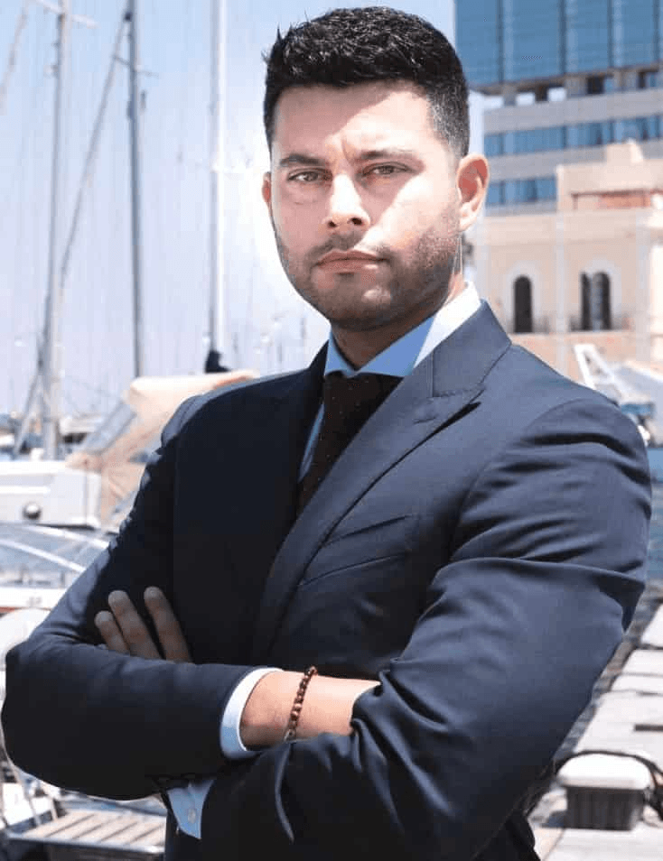 A confident man in a business suit stands with his arms crossed at a marina.