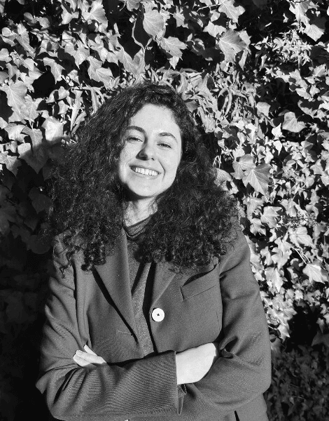 A black and white photo of a smiling woman with curly hair, standing in front of a leafy background.