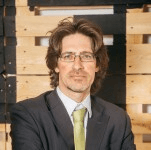 A professional man with medium-length hair wearing glasses, a dark suit, and a green tie stands confidently in an office setting with wooden pallet background.
