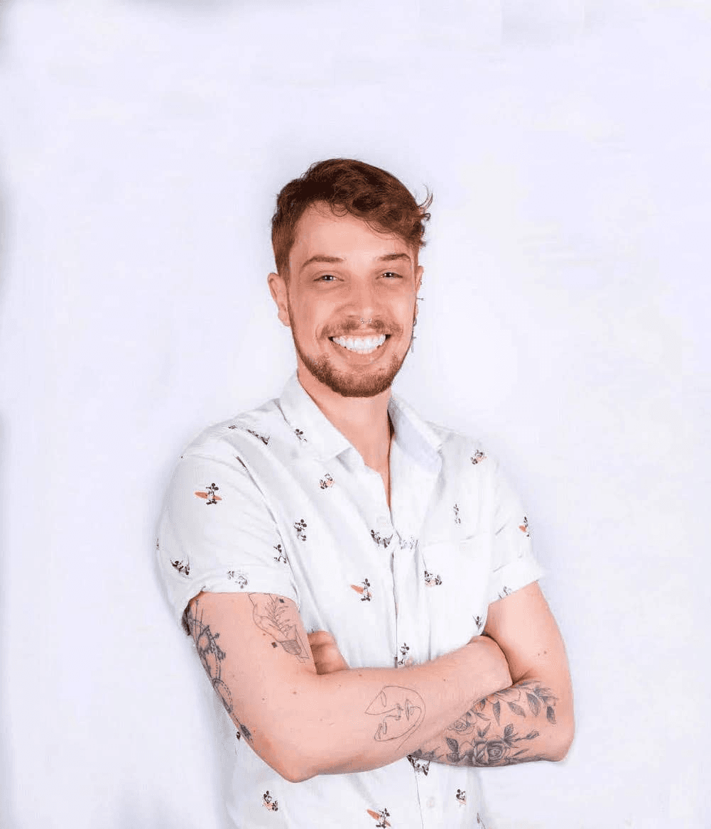 A young man with a beard, tattoos, and a floral shirt smiling confidently at the camera.