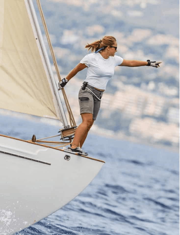 A woman balances on the edge of a sailing boat, extending her arms for stability.