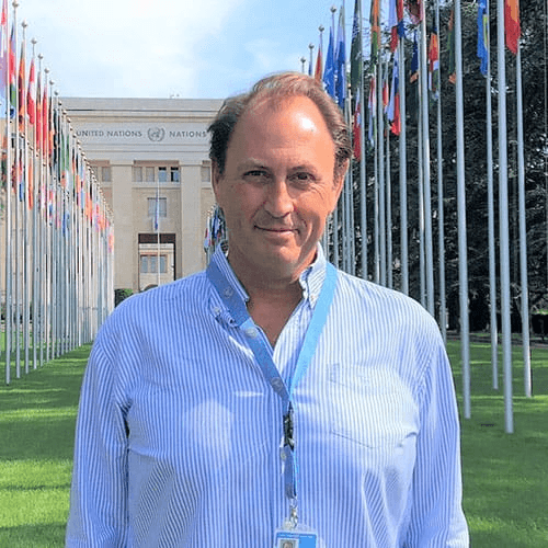 A man stands in front of the United Nations building, wearing a blue striped shirt and a badge.