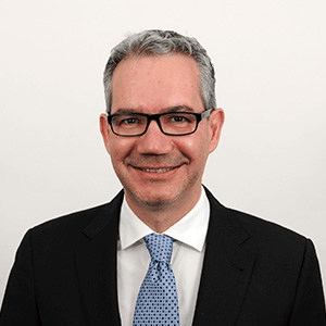 A professional portrait of a smiling man wearing glasses, a black suit, white shirt, and blue tie.