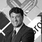 A grayscale image of a smiling man in a suit and tie standing in front of a logo.