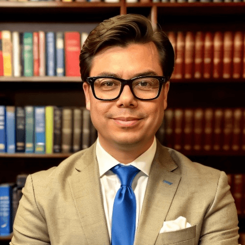 A professional man wearing glasses, a beige suit, and a blue tie is smiling in front of a bookshelf.