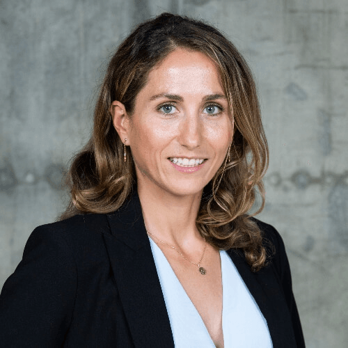 A professional portrait of a woman with long brown hair wearing a black blazer and white top, smiling against a gray background.