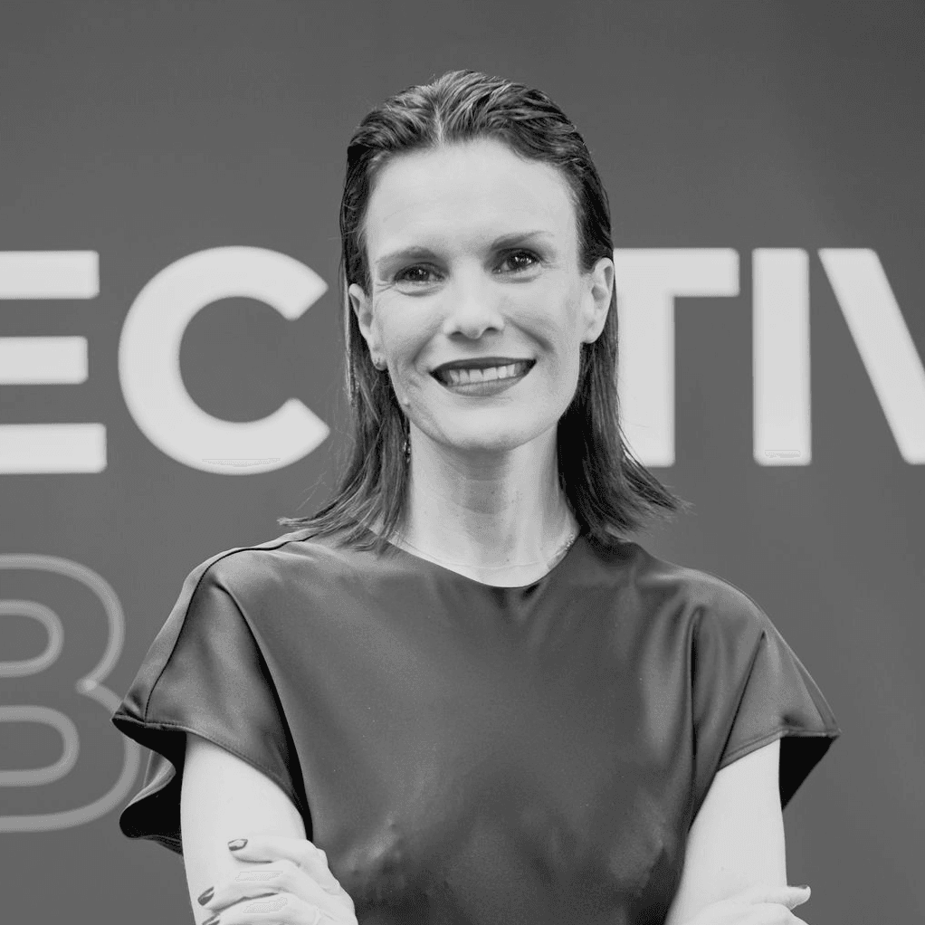 A black and white photo of a smiling woman with long hair, standing with her arms crossed in front of a sign that reads 'EFFECTIVE'.