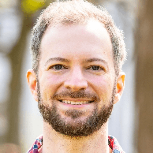 A smiling man with a beard and a plaid shirt standing outdoors.