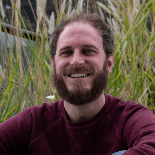 A smiling man with a beard sitting outdoors with tall grass in the background.