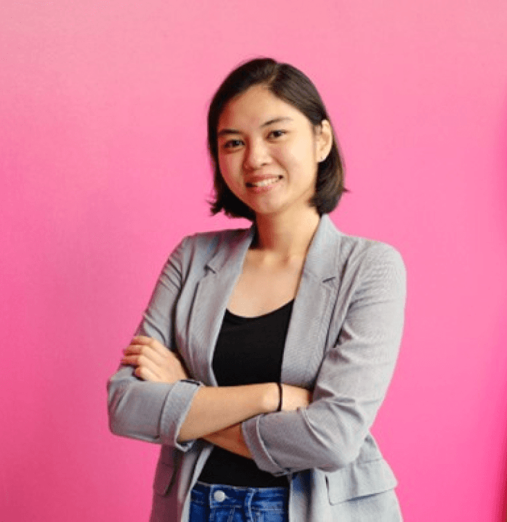 A young woman in a gray blazer and jeans stands smiling with her arms crossed against a pink background.