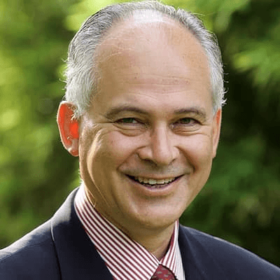 A portrait of a smiling middle-aged man wearing a suit and a red striped tie, set against a blurred green background.