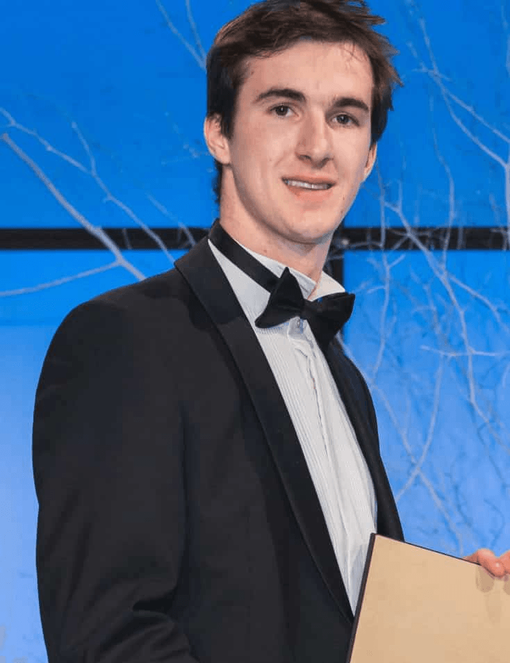A young man in a tuxedo holding a certificate at an event with a blue background.